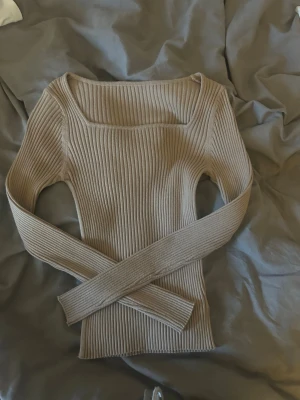Beige ribbstickad topp med fyrkantig ringning - Trendig beige ribbstickad topp med lång ärm och snygg fyrkantig ringning. Figurnära passform och stretchigt material som ger en modern look. Perfekt att styla med jeans eller kjol för en clean outfit.