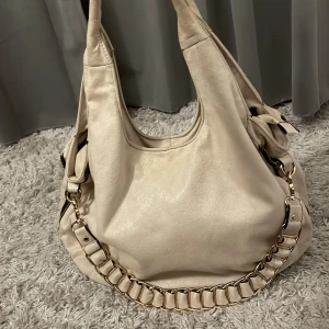 Beige axelväska med kedjedetalj - Snygg stor beige axelväska med guldfärgad kedjedetalj på axelremmen. Väskan har en dragkedja på baksidan, flera fack inuti för enkel organisering och rymmer mycket. Perfekt för att bära med sig allt man behöver under dagen.