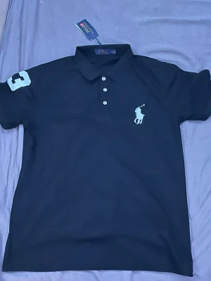 Svart pikétröja från Polo Ralph Lauren - Klassisk svart pikétröja från Polo Ralph Lauren med vit logga på bröstet och siffran 3 på ärmen. Tröjan har krage, tre knappar framtill och korta ärmar. Perfekt för en stilren och sportig look.