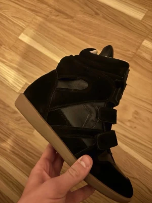 Isabel marant skor - Snygga svarta sneakers med högt skaft och tre kardborreband. Skorna har en mix av mocka och läderdetaljer samt en klassisk gummisula i beige. Perfekt för dig som gillar streetstyle och vill ha något unikt på fötterna.