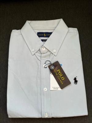Ljusblå skjorta från Polo Ralph Lauren - Klassisk ljusblå skjorta från Polo Ralph Lauren med button-down krage och broderad svart logga på bröstet. Skjortan har vita knappar och är tillverkad i mjuk bomull. Perfekt för en clean och stilren look.