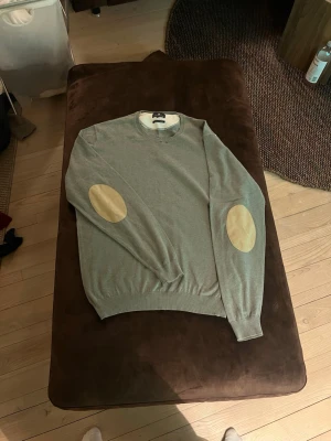 Grå cashmere-tröja Hansen & Jacob - Snygg grå stickad tröja från Hansen & Jacob i mjuk cashmere. Tröjan har rund halsringning och beiga mockadetaljer på armbågarna för en klassisk look. Perfekt för dig som gillar stilrena och tidlösa plagg.