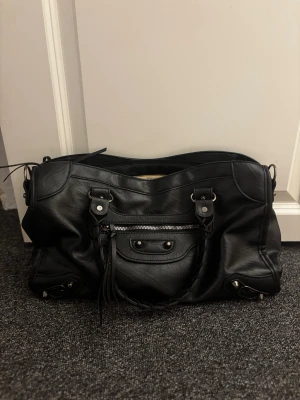 Svart City handväska - Från Designbyamore Säljer en klassisk svart City handväska i mjukt läder med silverfärgade metalldetaljer och dragkedja framtill. Väskan har dubbla handtag med flätad detalj och dekorativa nitar. Perfekt för dig som gillar ikoniska designeraccessoarer.