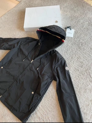 Svart vindjacka från Moncler - Snygg svart vindjacka från Moncler med huva och dragkedja framtill. Jackan har två fickor med tryckknappar och Moncler-logga på ärmen. Insidan av huvan har en röd, vit och blå rand. Tillverkad i lätt, vattenavvisande material.