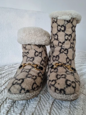 Beige fårskinnstofflor från Gucci - Säljer ett par beige tofflor från Gucci med GG-monogram i svart och guldfärgade detaljer framtill. Tofflorna har mjukt fårskinnsfoder och en högre skaftdesign som kan vikas ner. Perfekta för dig som vill ha både lyx och komfort hemma.