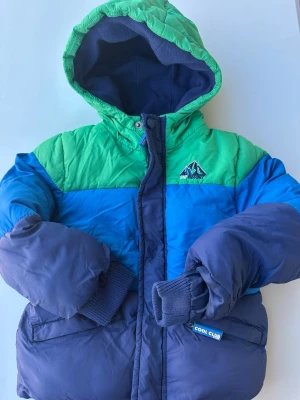 Färgglad dunjacka från Cool Club - Snygg dunjacka i blått, grönt och mörkblått från Cool Club. Jackan har huva, dragkedja och två fickor framtill. Perfekt för kalla vinterdagar och har en sportig look med broderad logga på bröstet.litet hål finns vid arm se bild 