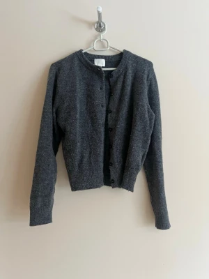 Mörkgrå cropped kofta - Mysig mörkgrå kofta med rund halsringning och knappar framtill. Modellen är cropped och har långa ärmar samt ribbade muddar vid ärmslut och nederkant. Perfekt att slänga över en topp för en enkel och stilren look.