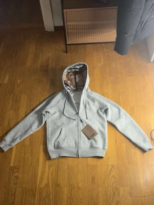 Grå zip hoodie med mönstrat foder - Snygg grå hoodie med dragkedja från Burberry. Hoodien har en klassisk huva med det ikoniska rutiga fodret, två fickor fram och ribbade muddar. Perfekt för en avslappnad och stilren look. Materialet känns mjukt och bekvämt. Passar S med 😊
