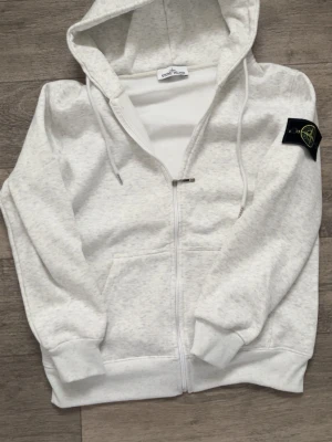 Stone island zip hoodie  - En grå Stone Island zip-hoodie med ett mjukt och skönt innermaterial som håller dig varm.   Storlek M och passar perfekt för kallare väder eller som en stilren vardagströja.    Hör gärna av dig vid minsta fundering!