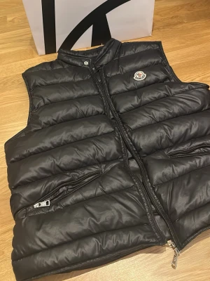 Svart dunväst från Moncler - Snygg svart dunväst från Moncler med quiltad design och hög krage. Västen har dragkedja framtill, två sidofickor med dragkedja och Moncler-logga på bröstet. Perfekt för lager-på-lager och streetstyle. Materialet är glansigt och känns lyxigt. Storlek M. Skick 8/10. Mitt pris 1800kr vid snabbaffär kan gå ner något 100kr. Skriv vid frågor eller intresse!
