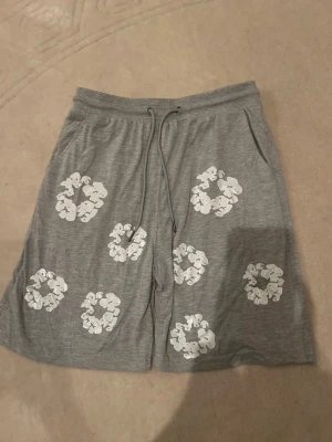Grå denim tears shorts - Grå shorts med vita blommönster och snörning i midjan. Tillverkade i mjukt bomullsmaterial och har en avslappnad passform. Perfekta för varma dagar och chill stunder.