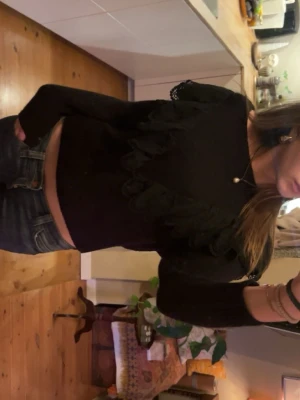 Svart tröja med volangdetaljer - Trendig svart tröja med volangdetaljer framtill och på ärmarna. Långärmad modell i mjukt material som passar perfekt till jeans eller kjol. Enkel men ändå unik design som ger outfiten det lilla extra.