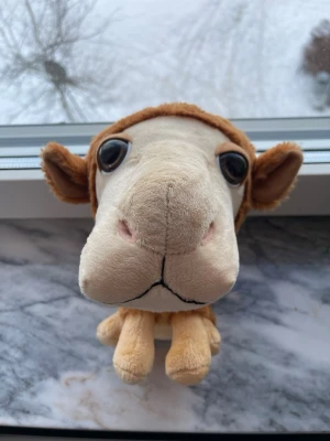 Mjukisdjur kamel med stora ögon - Söt mjukisdjur i form av en kamel med stora svarta ögon och mjuk, lurvig brun päls på ryggen. Kroppen är beige och kamelen har fyra ben samt små öron. Perfekt som gosedjur eller dekoration i rummet.