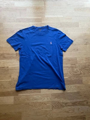 Blå t-shirt från Polo Ralph Lauren - Klassisk blå t-shirt från Polo Ralph Lauren med rund halsringning och korta ärmar. På bröstet finns den ikoniska rosa broderade loggan. Tillverkad i mjuk bomull för en skön känsla och enkel stil.