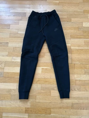 Svarta mjukisbyxor från Nike - Säljer ett par svarta mjukisbyxor från Nike med dragsko i midjan och ribbade muddar vid bensluten. Byxorna har en slim fit passform och diskret Nike-logga på vänster lår. Storlek XS, ny skick