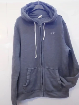 Grå zip hoodie från Hollister - Mysig grå hoodie från Hollister med dragkedja, vita snören och broderad logga på bröstet. Hoodien har två stora fickor framtill och en klassisk huva. Tillverkad i mjukt bomullsmaterial som är perfekt för chill dagar.
