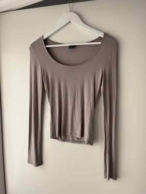 Beige långärmad topp Gina Tricot  - Säljer en enkel och stilren beige långärmad topp från Gina Tricot. Toppen har en rund, djup urringning och är gjord i mjukt, stretchigt material som sitter snyggt på kroppen. Perfekt att matcha med jeans eller kjol för en clean look.