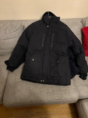 Svart parkasjacka med fickor - Snygg svart parkasjacka med hög krage och dragkedja framtill. Jackan har två stora fickor med lock och knappar samt två bröstfickor med dragkedja. Perfekt för kalla dagar och har en klassisk, stilren look.