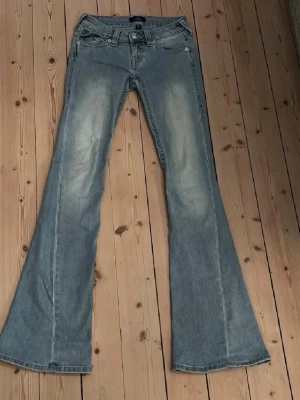 Blå bootcut jeans från True religion  - Säljer ett par ljusblå bootcut jeans från True Religion med slitningar framtill och bakfickor med lock och knappar. Jeansen har låg midja och klassisk femficksdesign. Perfekta för dig som gillar retrostil och vill ha ett par riktigt snygga jeans!