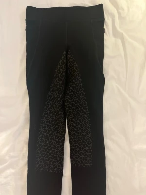 Svarta ridbyxor från Mountain Horse - Säljer ett par svarta ridbyxor från Mountain Horse med geometriskt mönstrade gripzoner på insidan av benen och bak. Byxorna har hög midja och stretchigt material som ger bra rörelsefrihet. Perfekta för träning i stallet. 🐴💞
