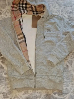 Grå zip-hoodie från Burberry - Snygg grå zip-hoodie från Burberry med klassiskt rutigt foder i huvan. Hoodien har dragkedja framtill, två fickor och ribbade muddar vid ärmar och nederkant. Tillverkad i mjukt bomullsmaterial och har en diskret logga på bröstet. Perfekt för en avslappnad och trendig look.
