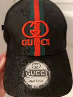 Svart Gucci keps med röd-gröna detaljer - Snygg svart snapback-keps från Gucci med klassiska röd-gröna ränder och broderad logga framtill. Kepsen har diskret GG-mönster i tyget och justerbar baksida. Insidan är fodrad med Gucci-tryck. Perfekt för dig som vill ha en exklusiv och trendig accessoar.