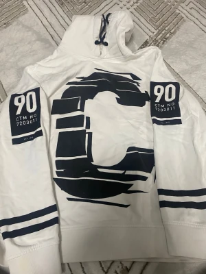 Vit hoodie med svart tryck - Snygg vit hoodie med marinblå grafiskt tryck på bröstet och armarna. Stora siffror '90' och text på båda ärmarna samt två marinblå ränder vid ärmsluten. Huva med dragsko och avslappnad passform. Perfekt för en cool och sportig stil. Lite smuts men tror kan tvättas bort. 