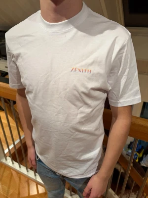 Vit t-shirt från Zenith - Snygg vit t-shirt från Zenith med diskret logga i regnbågsfärger på bröstet. Klassisk passform med rund hals och korta ärmar. Tillverkad i mjuk bomull som känns skön mot huden. Perfekt till jeans eller shorts för en clean och enkel look. Tröjan finns i storlek XS, S och M