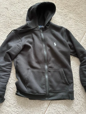 Svart hoodie från Polo Ralph Lauren - Svart hoodie med dragkedja från Polo Ralph Lauren. Klassisk design med huva, två fickor framtill och den ikoniska vita polospelaren broderad på bröstet. Tillverkad i mjukt bomullsmaterial som är skönt att bära. Perfekt för en avslappnad och stilren look.