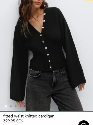 Svart ribbad kofta med knappar - Trendig svart kofta med ribbad midja och vida ärmar. Koftan har djup V-ringning och vita knappar längs hela framsidan. Stickad i mjukt material som ger en snygg passform och modern look.