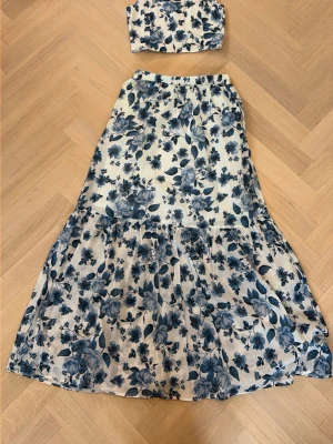 Blommig långkjol i blått och vitt - Säljer en flowy långkjol med blått blommönster på vit botten med en matchande topp till. Kjolen har resår i midjan och volangdetalj nedtill för en extra luftig känsla. Kjolen är högmidjad/medelmidjad, men kan ha den som lågmidjad. Har använt dem 2 gånger.