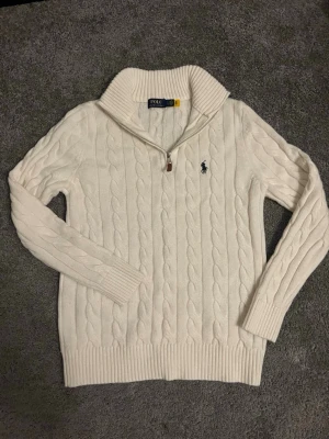 Vit kabelstickad tröja Polo Ralph Lauren - Kabelstickad vit tröja från Polo Ralph Lauren med halv dragkedja och hög krage. Tröjan har det klassiska broderade logotypen på bröstet och ribbstickade muddar. Perfekt för lager-på-lager och snygg till jeans.