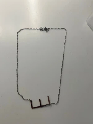 Silvrigt halsband med minimalistiskt hänge - Snyggt halsband i silverfärgad metall med ett unikt, geometriskt hänge i form av raka linjer. Kedjan är tunn och har ett klassiskt lås. Perfekt för dig som gillar enkel och modern design.      Aldrig använt 