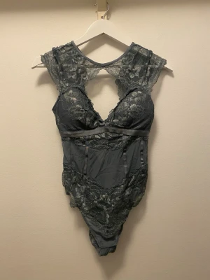 🩶 Mörkgrå/Silver Spetsbody - ✨ Snygg mörkgrå-silver body i spets med vackra blommönster och öppen rygg. Modellen har korta ärmar och djup urringning framtill. Tillverkad i genomskinligt spetsmaterial som ger en elegant och feminin look. Använd en gång. Från Hunkemöller. Storlek M. ✨✨ 