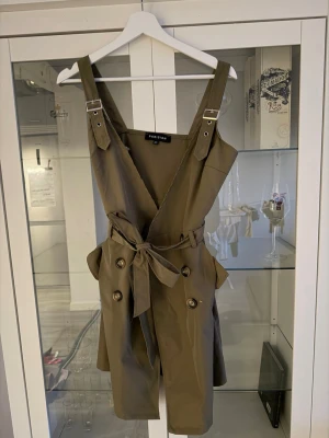 Khakigrön skort från Parisian - Supersnygg khakigrön skort från Parisian med trendiga knappar framtill, breda axelband med spännen och ett knytband i midjan. Skorten har en cool trenchcoat-inspirerad look och är gjord i ett mjukt, vävt material. Perfekt för dig som vill ha något unikt och stilrent.