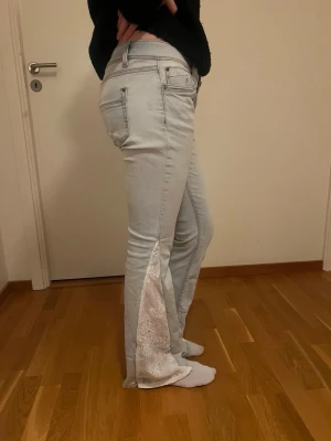 Ljusblå bootcut jeans med spetsdetalj - Unika ljusblå jeans med breda spetsinsatser längs bensluten. Jeansen har normal passform och klassisk femficksdesign. Spetsen ger en cool och personlig touch till looken. Perfekta för dig som vill sticka ut lite extra! Midjemåttet är 40 och innerbenslängden 79, storlek 36🩷