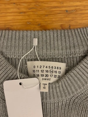 Grå stickad tröja från Maison Margiela - Snygg grå stickad tröja från Maison Margiela med rund halsringning och ribbstickade muddar. Tröjan har en klassisk passform och är tillverkad i ett mjukt material som passar perfekt till lager på lager. Diskret och stilren design.