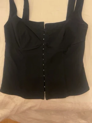 Svart korsett-topp med hyskor - Snygg svart korsett-topp med breda axelband och hyskknäppning framtill. Toppen har en figurnära passform och markerad byst. Perfekt att styla med jeans eller kjol för en trendig look.