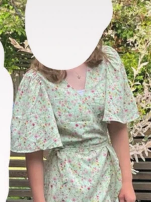 Grön blommig sommarklänning - Söt grön omlottklänning med blommigt mönster i rosa och vitt. Klänningen har korta, vida ärmar och knyts i midjan för en snygg passform. Perfekt för varma dagar och somriga tillfällen.