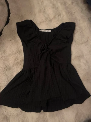 Svart topp med knyt  - Supersnygg svart topp v-ringning och knyt framtill. Toppen har volangdetaljer vid axlarna och är lätt utsvängd nedtill. 