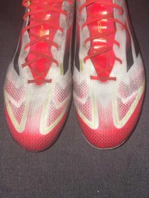 Adidas F50 Elite AG  - Säljer ett par Adidas F50 fotbollsskor i vitt med röda och gula detaljer samt svarta ränder. Skorna har snörning, platt sula med dobbar och är tillverkade i syntetmaterial. Kommer med matchande röd F50-påse och extra innersulor. Perfekta för dig som vill sticka ut på planen. Skorna är använda i 2-3 månader. pruta gärna!!! Köpta för 2419kr(unisport)