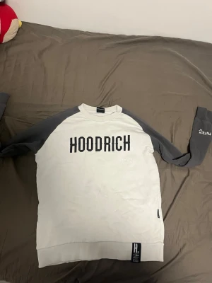 Vit och grå sweatshirt från Hoodrich - Säljer en snygg sweatshirt från Hoodrich med vit kropp och mörkgrå ärmar. Tröjan har tryckt HOODRICH-logga på bröstet och textdetaljer på ryggen. Klassisk rund halsringning och ribbade muddar. Perfekt för en streetwear-look.