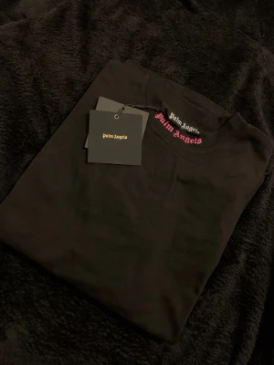 Svart långärmad topp Palm Angels - Svart långärmad topp från Palm Angels med stor rosa logga i gotisk stil över axlarna. Tillverkad i mjuk bomull och har en klassisk rund halsringning. Perfekt för dig som gillar streetwear och vill sticka ut med en ikonisk detalj.