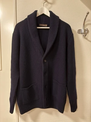 Oscar Jacobsson Roland Cardigan  - Nypris 3000 kr. Snygg mörkblå ribbstickad kofta från Oscar Jacobsson med sjalkrage och knäppning framtill. Koftan har två stora fickor och långa ärmar. Perfekt för lager på lager och en stilren look.