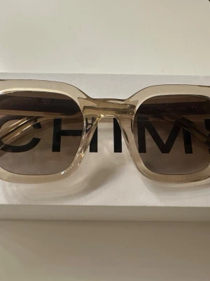 Beige CHIMI solglasögon fyrkantiga - Snygga solglasögon från CHIMI med transparent beige båge och fyrkantig form. Glasen är mörka och ger en cool kontrast till den ljusa ramen. Perfekta för dig som vill ha en modern och trendig look. Designade i Stockholm. I färgen ecru och modelen 04