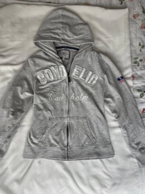 Grå hoodie från Bondelid med tryck - Säljer en grå hoodie från Bondelid med broderat vitt tryck 'Bondelid Stockholm 1956' på bröstet och amerikansk flagga på ärmen. Hoodien har dragkedja, huva och en stor magficka. Perfekt för chill dagar och snygg streetstyle.