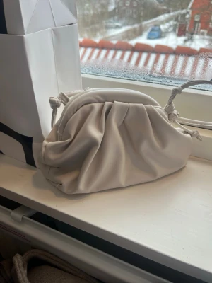 Vit axelremsväska med dragsko - Trendig vit handväska i mjukt, slätt material med dragsko och axelrem. Väskan har en puffig, veckad design som ger en modern look. Perfekt för dig som gillar stilrena accessoarer med det lilla extra.