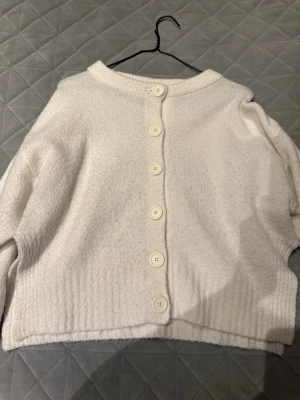 Vit kofta med knappar från 157 - Mysig vit kofta från 157 med stora matchande knappar framtill. Koftan har en rak passform, ribbade muddar och rund halsringning. Perfekt för lager på lager och enkel att styla till olika outfits.