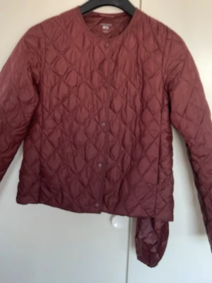 Vinröd  quiltad pufferjacka Uniqlo - Säljer en brun quiltad pufferjacka från Uniqlo i storlek S. Jackan har rund halsringning, tryckknappar framtill och ett lätt vadderat material som håller dig varm. Perfekt för kalla dagar och enkel att matcha med olika outfits.