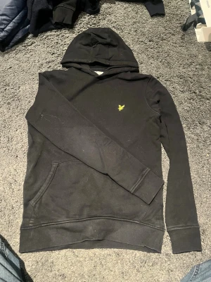 Svart hoodie från Lyle & Scott - Säljer en svart hoodie från Lyle & Scott med klassisk gul logga på bröstet. Hoodien har magficka, ribbade muddar och huva. Tillverkad i mjukt bomullsmaterial och har en clean, stilren look som passar perfekt till jeans eller joggers.